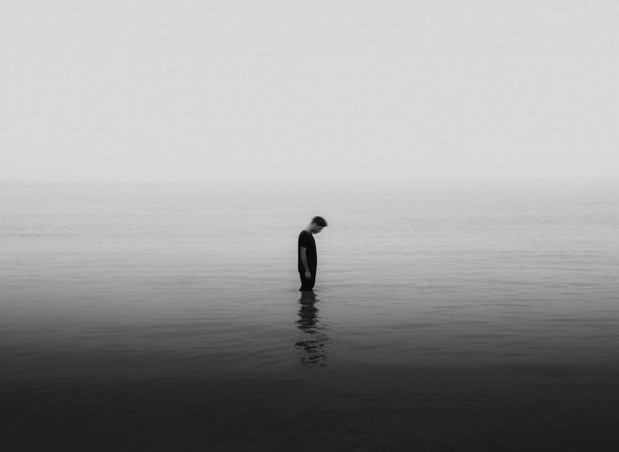 A black and white minimalist image showing a lone person standing waist-deep in a vast, misty body of water.