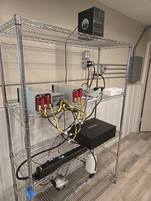 A metal wire shelving unit holds a small-scale DigiByte mining rig, including power supplies with visible "DigiByte" branding, a miner unit, and an iPollo device, alongside an electrical panel and mounted power outlet. The setup reflects an in-progress home crypto mining operation as referenced in the caption.