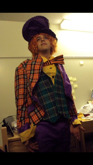 A person in a Mad Hatter costume featuring a purple top hat, orange wig, yellow bow tie, plaid jacket, and purple pants, standing in a classroom setting.