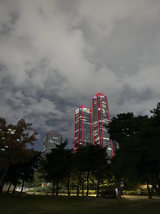 A nighttime photograph of a park with trees in the foreground and two tall skyscrapers illuminated with red lighting against a cloudy sky.
