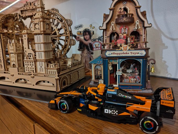 A McLaren F1 model car sponsored by OKX and Chrome sits displayed on a wooden shelf next to a "Happyduke Toys" storefront model and a wooden Ferris wheel kit, with the OKX logo visible on both the car and a circular sign in the background, showcasing a thoughtfully gifted building kit that has been assembled and added to the recipient's collection.