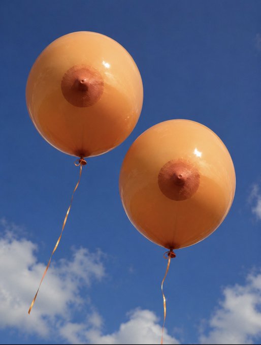 Two orange balloons with nipple designs float against a blue sky with white clouds.
