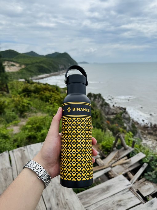 A hand wearing a silver watch holds a black and yellow Binance-branded water bottle against a coastal landscape with mountains and ocean.