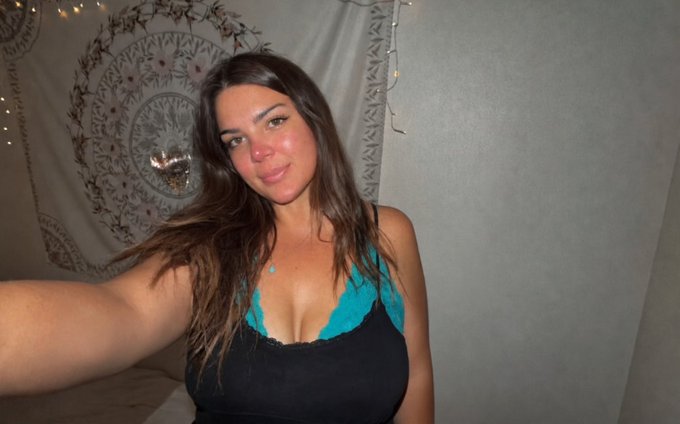 A young woman with long dark hair takes a selfie indoors wearing a teal bikini top under a black tank top, with a mandala tapestry and string lights in the background.