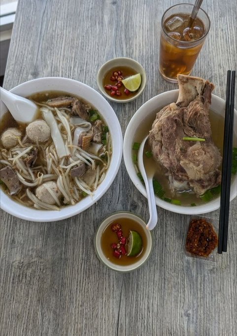 A spread of Vietnamese-style noodle soup with beef, bean sprouts, meatballs, and a large bone-in meat dish, accompanied by iced tea, lime-chili dipping sauces, and chili paste, arranged on a wooden table — a hearty "good morning CT" breakfast that invites reflection on how life's simple pleasures, like a warm bowl of soup, make living worthwhile.