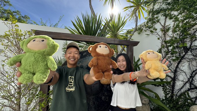 Two people smiling and holding up three plush monkey toys in green, brown, and tan colors, standing outdoors under a wooden structure surrounded by tropical plants.