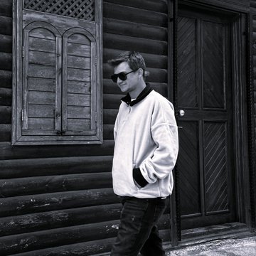 A black-and-white photo shows a man wearing sunglasses and a light jacket standing in front of a wooden log cabin with a paneled door and shuttered windows.