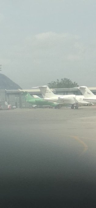 The image shows an airport tarmac with several airplanes parked, including one green airplane and two white planes. A person can be seen standing near the airplanes, possibly a worker or a passenger. The scene conveys a sense of organization and efficiency at the airport, as all the airplanes are neatly parked in their designated areas.