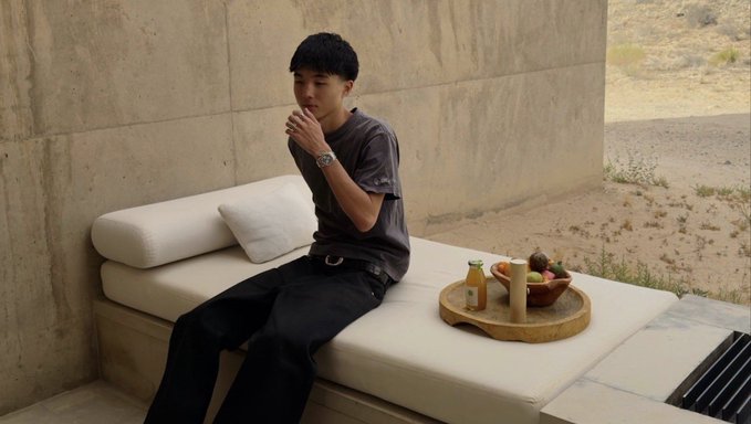 A young man sits relaxed on a cushioned outdoor daybed in a desert setting, sipping a drink with eyes closed in a serene moment, while a tray of fresh fruit and a beverage rests beside him—evoking a "good morning" vibe of leisure and prosperity that aligns with the optimistic "road to $40m" sentiment of the post.