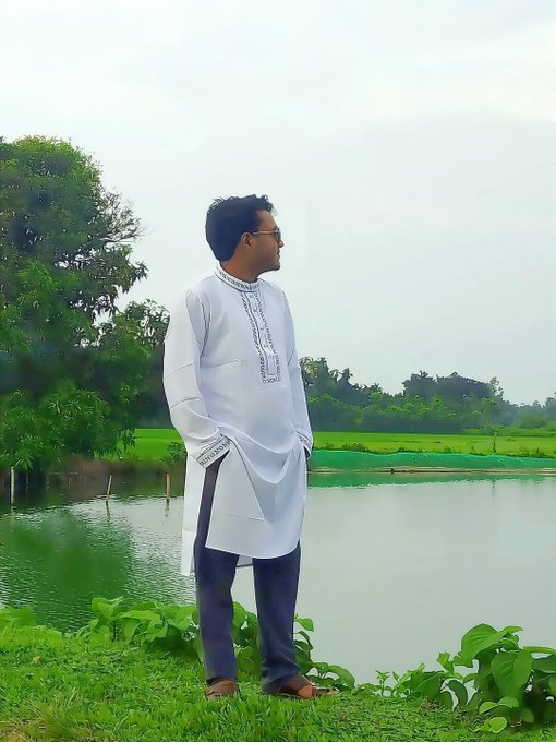 A man wearing glasses and a white embroidered kurta stands outdoors on a grassy bank next to a pond, with green fields and trees in the background.