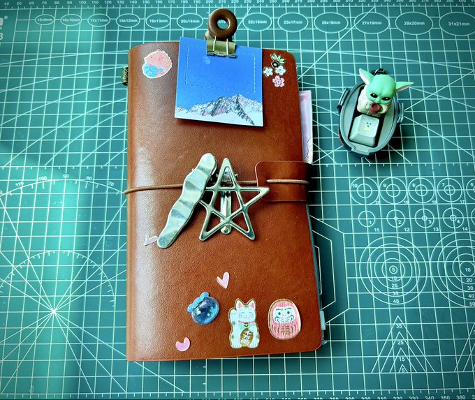 A brown leather traveler's notebook decorated with enamel pins and charms sits on a green cutting mat next to a small figurine of Grogu and a car key fob.