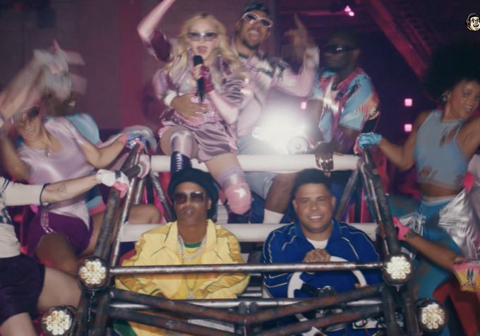 A group of people dressed in flashy, retro outfits—including one person in a yellow puffer vest and beanie and another in a blue jacket—are riding together in a rugged vehicle, with a blonde figure holding a microphone in the center. The party-like, over-the-top scene visually captures the post's playful "gang back together" sentiment, evoking the excitement and chaos of a crypto bull market.