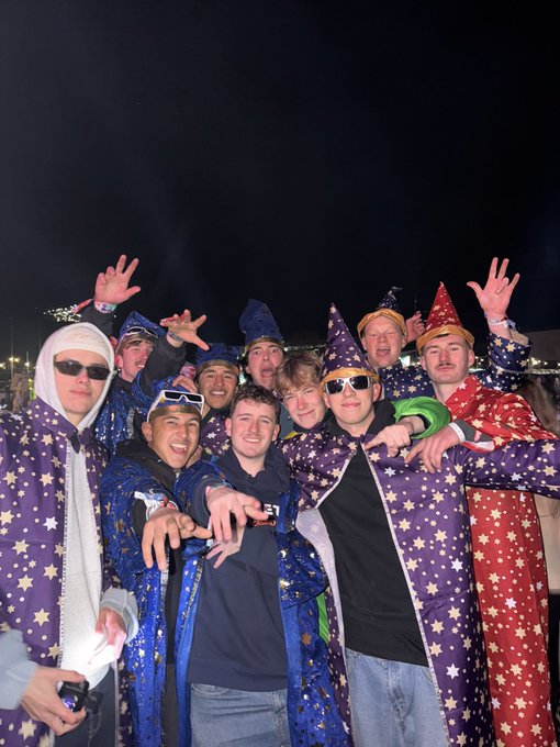 A group photo of young men at night wearing starry wizard robes and pointed hats, posing enthusiastically with hand gestures for the camera.