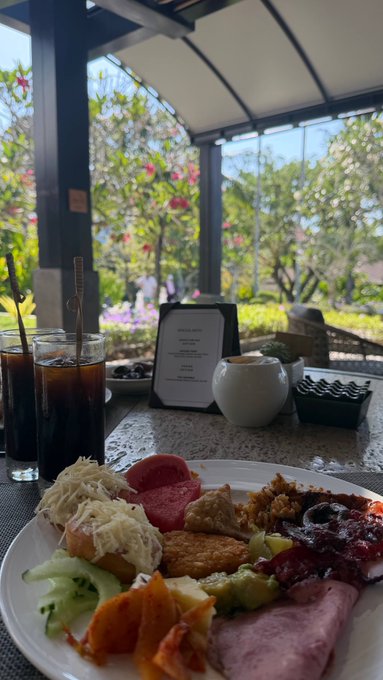 A breakfast spread sits on a table, featuring a plate with fried eggs, sausages, cucumber, papaya, and other items, alongside iced coffee in glasses and a small white teapot, with a lush garden view in the background.