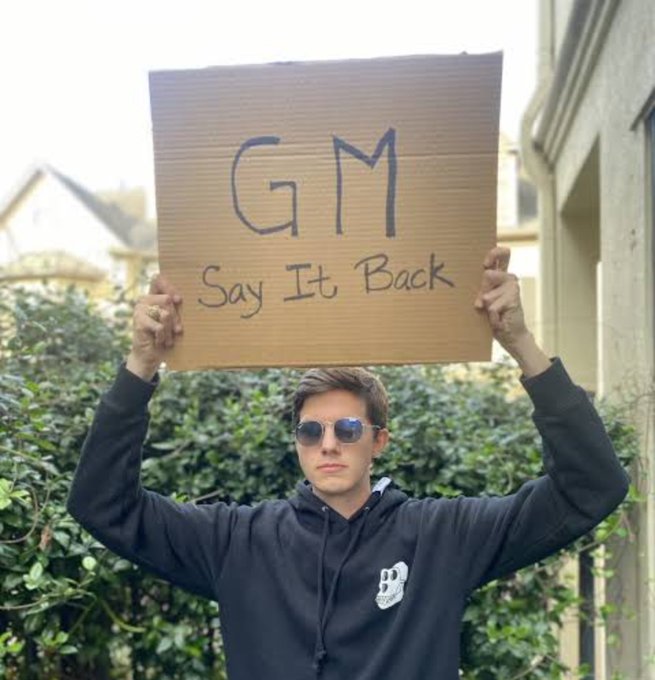 A young man wearing sunglasses and a black hoodie stands outdoors holding up a cardboard sign that reads 'GM Say It Back' above his head.