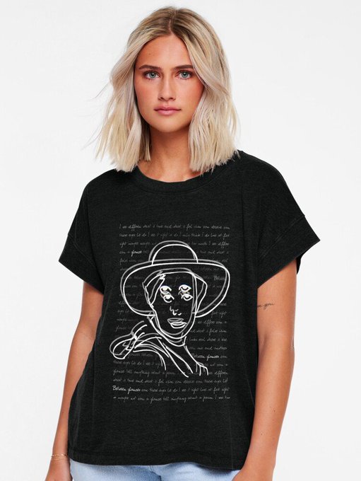 A woman wearing a black t-shirt featuring a white line-drawing illustration of a face wearing a hat surrounded by handwritten-style text.