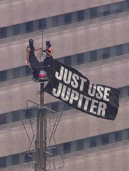 Two protesters scale a communications tower near a large building, unfurling a black banner that reads "JUST USE JUPITER" in bold white letters, echoing the post's slogan.