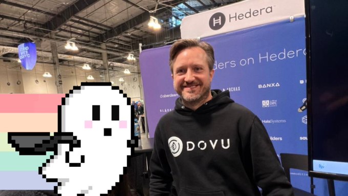 A man smiles while wearing a black 'DOVU' hoodie at an event with a 'Hedera' banner, next to a pixelated ghost graphic with a pride flag background.