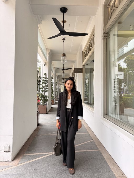 A woman wearing a black blazer, white top, black trousers, and carrying a handbag stands in a covered walkway with white columns, hanging fans, and large windows.