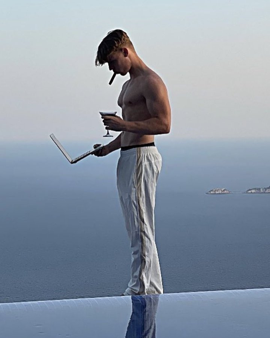 The image features a shirtless man standing at the edge of a pool, holding a laptop computer while smoking a cigar. The man appears to be enjoying his time by the water with his drink. The photo captures an interesting juxtaposition between technology use and leisure activities, as well as the man's casual attire in contrast to the formal setting of using a laptop outdoors.