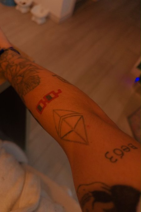 A person's forearm displaying multiple tattoos including a geometric cube/diamond shape, the number "863," and other designs, photographed in warm lighting.