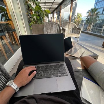 A man with a watch works on a laptop outdoors at a patio setting, with another open laptop beside him and palm trees in the background. The caption "Ăn có Tập có Làm có Build X" suggests a disciplined lifestyle of eating, training, and working, which is visually represented by the productive remote-work scene.