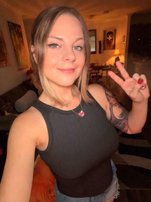 A selfie of a smiling woman with a nose ring and a 'PUMP' heart pendant necklace, making a peace sign with her tattooed arm in a cozy living room setting.