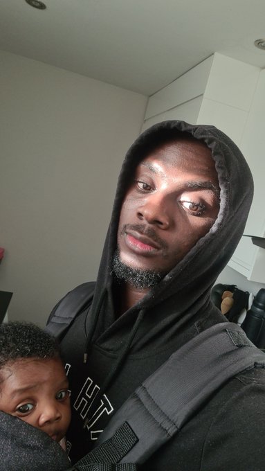 A man wearing a black hoodie looks into the camera while holding a young child close to his chest.