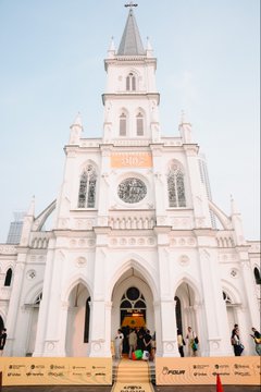 A grand white colonial-style church with a tall steeple serves as the backdrop for the BNB Super Meetup Singapore event, where attendees gather near the entrance, while promotional banners for "BNB" and "FractalVision" line the front steps.