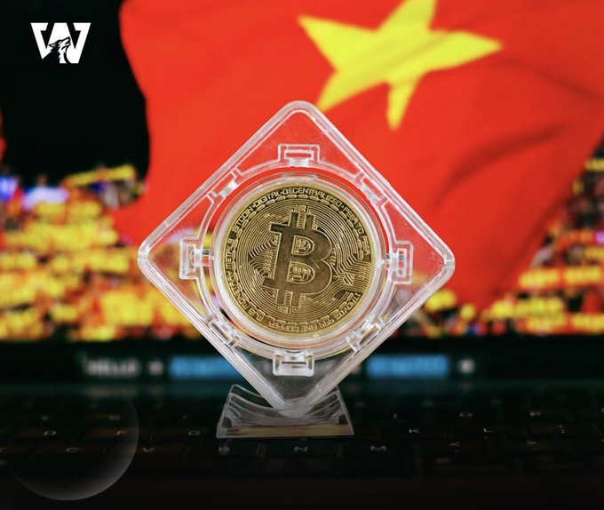 A physical gold-colored Bitcoin coin sits in a diamond-shaped clear plastic display case, placed on a laptop keyboard with a Vietnamese flag blurred in the background.