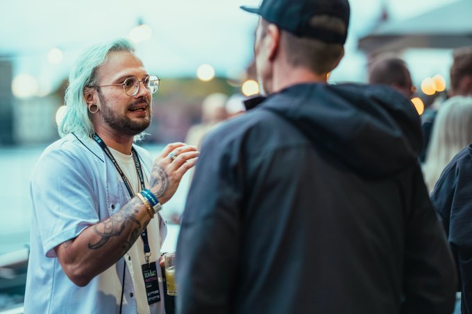A man with turquoise hair and glasses talks with another person at an outdoor event.