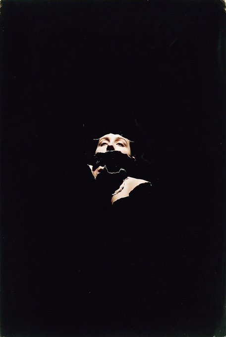 A dark, moody image showing a pale figure with an obscured lower face emerging from a black background, with only the eyes and forehead illuminated.