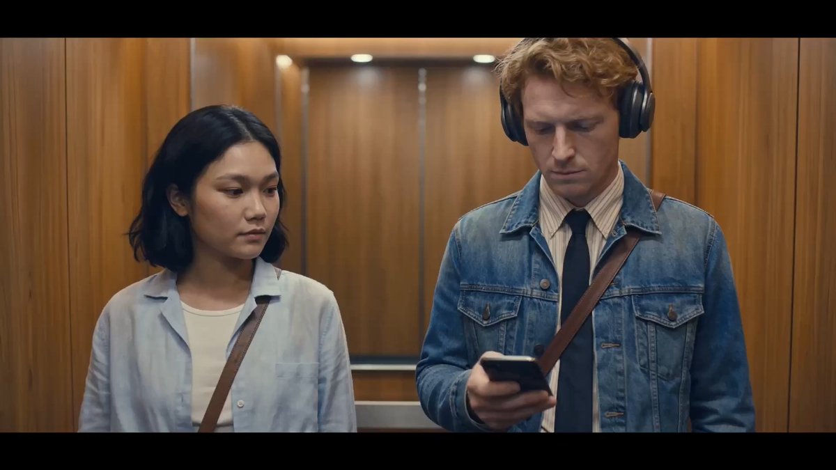 The image shows a man wearing headphones looking at his phone while standing next to a woman who is also holding her phone. The scene takes place inside an elevator, with a wooden panel visible behind them. There are two descriptions provided for this image: "Focus on what is in the image and how it relates to the post."