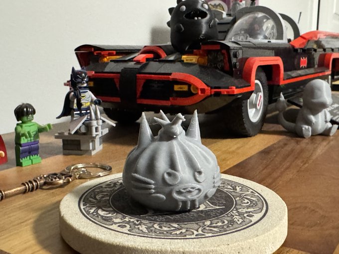 A 3D-printed gray cat dumpling character sits on a decorative coaster in front of the iconic 1989 Batmobile LEGO set, alongside a custom Batman minifigure and a Hulk figure, demonstrating the skill's ability to turn NFT artwork into physical figurines with just a few clicks.