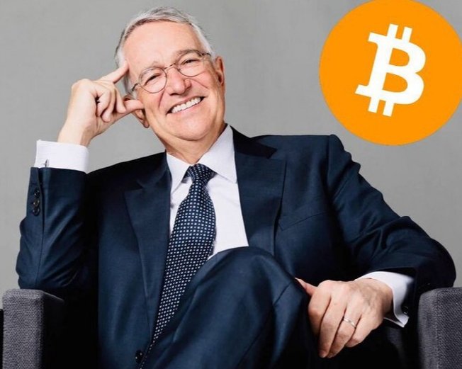 The image features a man dressed in a suit and tie, sitting comfortably in a chair with his hands resting on his lap. The man has a smile on his face as he looks directly at the camera. In front of him, there's an orange circle that says "bitcoin" written on it. The man is wearing glasses and appears to be older than 50 years old. He seems to be in a relaxed position while posing for the photo.