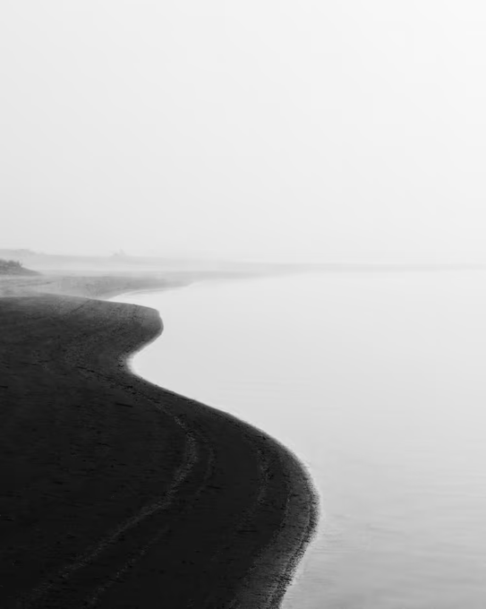 A black and white, minimalist photograph of a dark sandy shoreline curving into calm water with a foggy horizon.