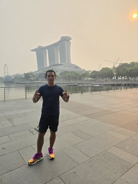 A man in a black t-shirt, black shorts, and bright yellow/orange running shoes stands on a paved plaza giving a double thumbs-up. The Marina Bay Sands hotel is visible in the background under a hazy sunset sky in Singapore.