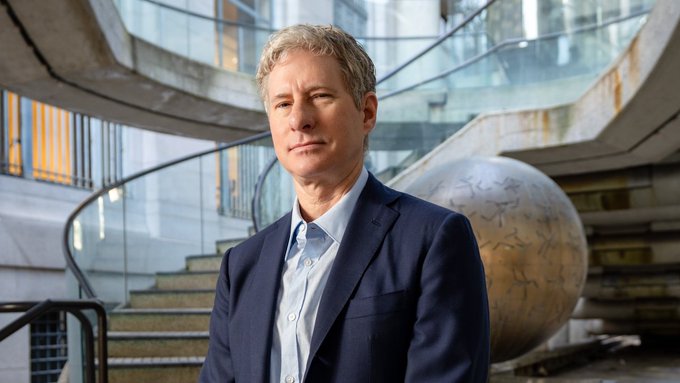 The image features a man dressed in a suit, standing next to a large globe or ball sculpture. The man appears to be posing for a photo while looking directly at the camera. In the background, there are stairs leading up to another level of the building. The caption above the image reads "CRAZY: Chris Larsen says 
Banks had decades to fix payments. They failed. $XRP did it in 10 years."". This text is related to a post about cryptocurrency and its impact on traditional banking systems, as suggested by the man's suit attire and the presence of the globe or ball sculpture.