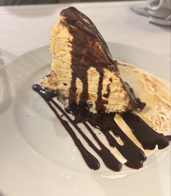 A slice of layered cake or cheesecake drizzled with chocolate sauce, served on a white plate alongside what appears to be a thin crepe or wafer.