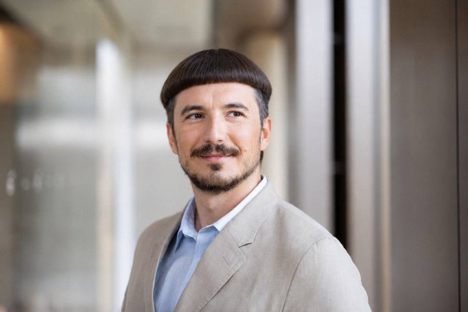 The caption: A smiling man with a distinctive bowl cut hairstyle and light beard poses in a beige blazer over a light blue shirt, fittingly showcasing the "Friar Tuck" hairdo referenced in the accompanying social media post.