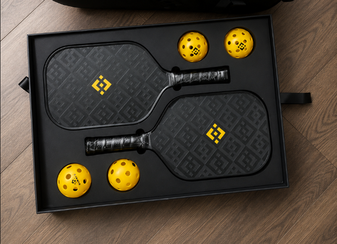 A black gift box containing a pickleball set, including two black and yellow patterned paddles branded with a logo, and four yellow perforated balls.