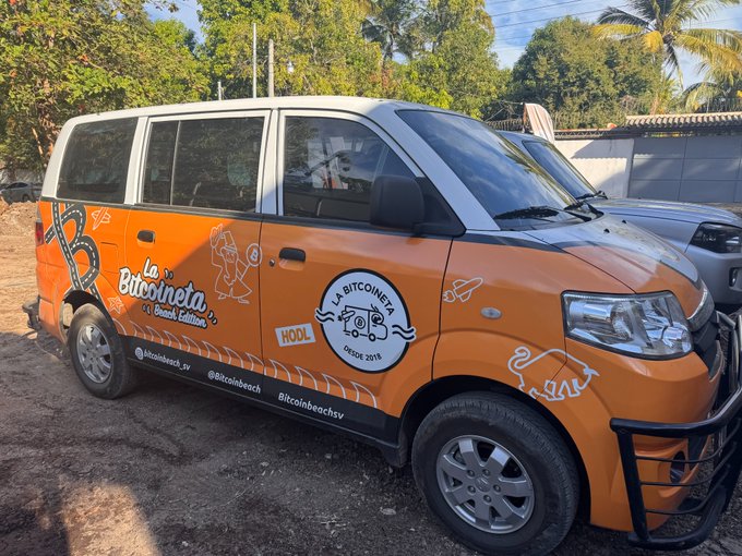 An orange van branded with 'La Bitcoineta' logos parked outdoors on a dirt surface near palm trees.
