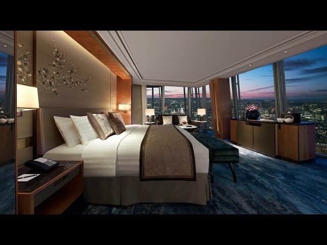 A luxurious hotel room with a large bed, neutral-toned décor, and floor-to-ceiling windows showcasing a city skyline at dusk.