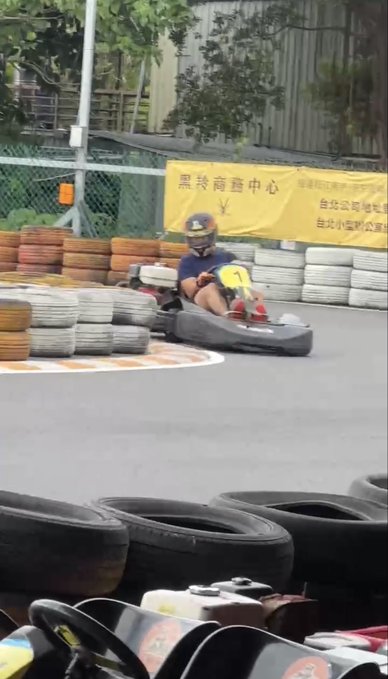 A person wearing a helmet drives a go-kart on a track lined with stacked tires, with a yellow banner displaying Chinese text in the background.