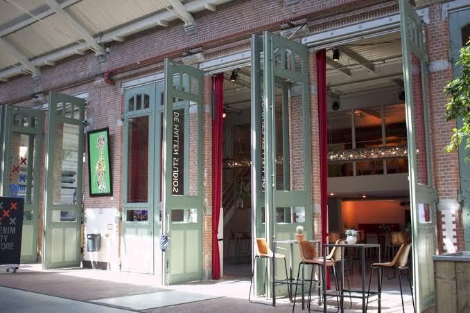 The exterior of a brick building featuring large green-framed glass doors with the text 'DE WINTHE LANDRO?' visible, showing an industrial-style interior space with red columns and modern tables and chairs.