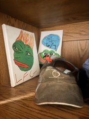 A green cap and two small canvas paintings (one featuring Pepe the Frog) sitting on a wooden floor.