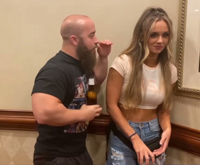 A bald, bearded man in a black graphic t-shirt stands next to a young woman with long blonde hair wearing a white crop top and denim skirt in an indoor setting.