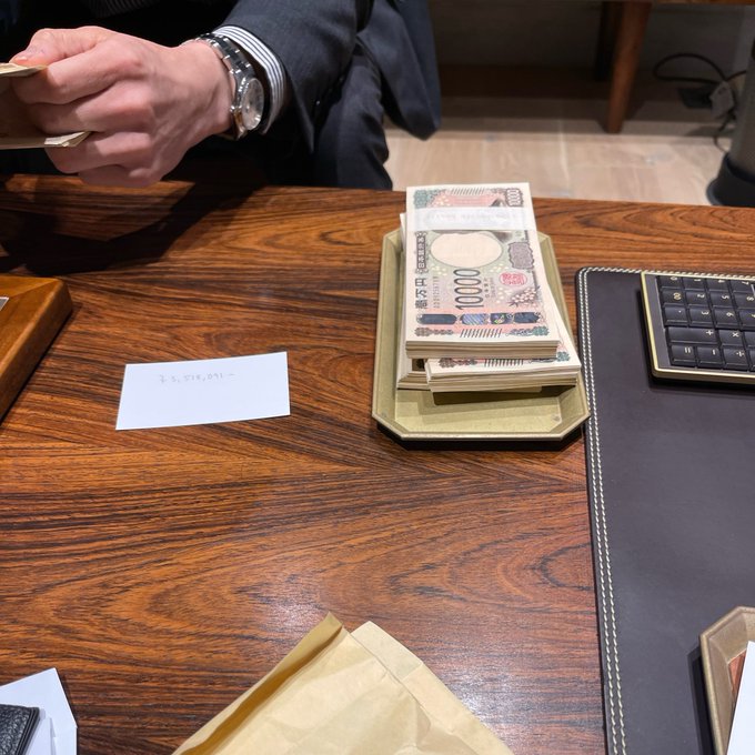A man in a suit sits at a wooden desk counting a stack of 10,000 Japanese yen bills in a tray, with a handwritten note reading "$3,556,081" visible on the table, seemingly showcasing the significant savings from switching to the fee-free @pumpfun trading app.
