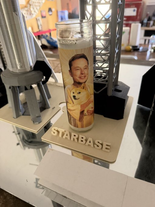 A candle featuring an illustration of Elon Musk holding the Doge dog sits on a 3D-printed "STARBASE" stand beside a model rocket, embodying the "Dogefather" moniker associated with Musk and the Dogecoin meme.