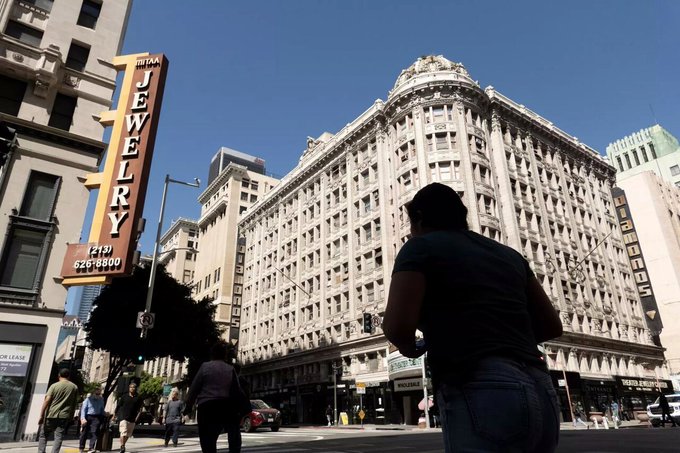 A street scene featuring the ornate white facade of a historic building and a vertical "JEWELRY" sign with a phone number 626-8800, with a person in the foreground and pedestrians on the sidewalk.